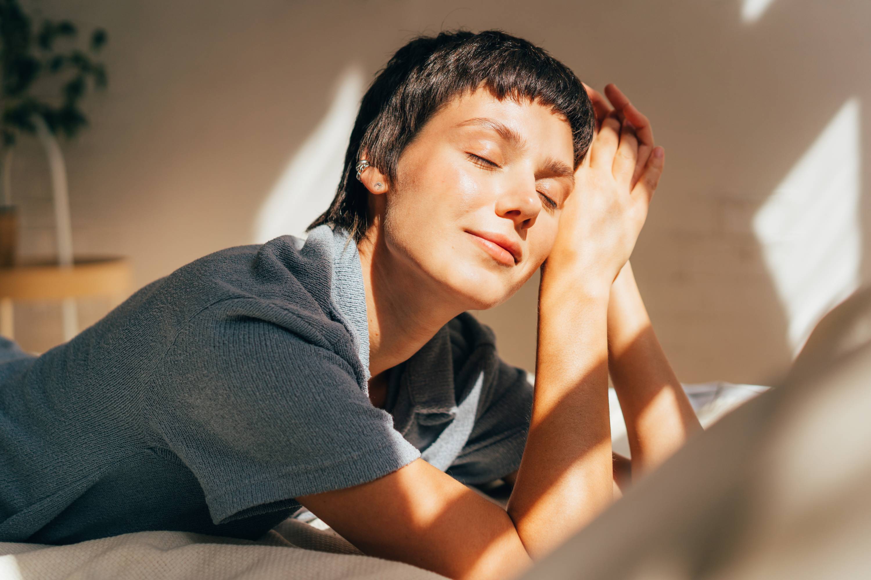 Eine Frau liegt ruhig und leicht lächelnd auf einem Bett in hellem, sonnigen Raum