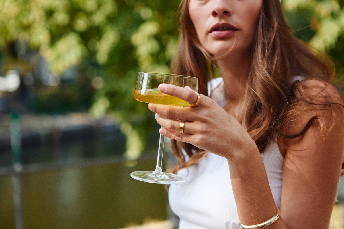 Junge Frau mit braunen Haaren und goldenem Schmuck hält ein Glas mit gelborangem Tee-Getränk