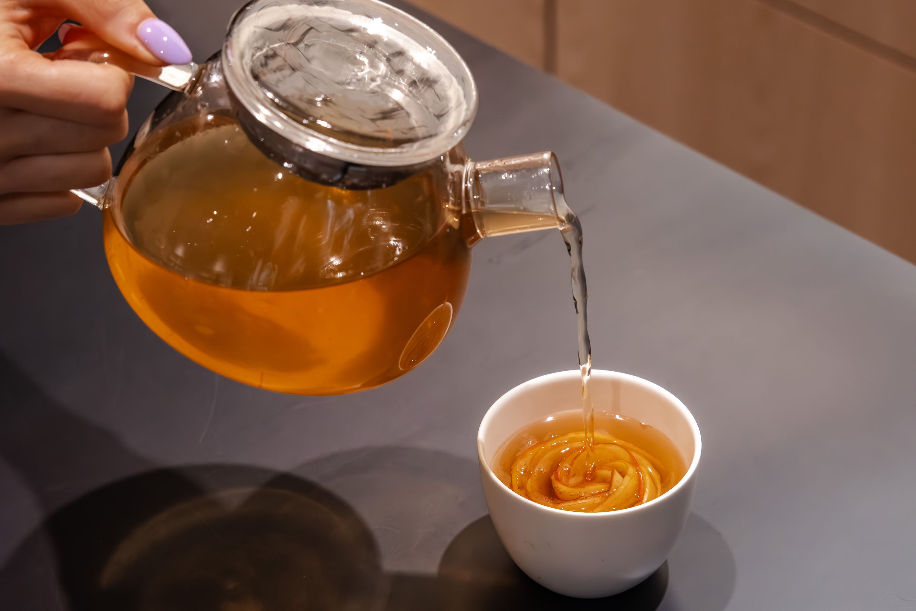 Hand mit lavendelfarbenem Nagellack schenkt Tee aus Glas-Teekanne in weiße Keramiktasse ein
