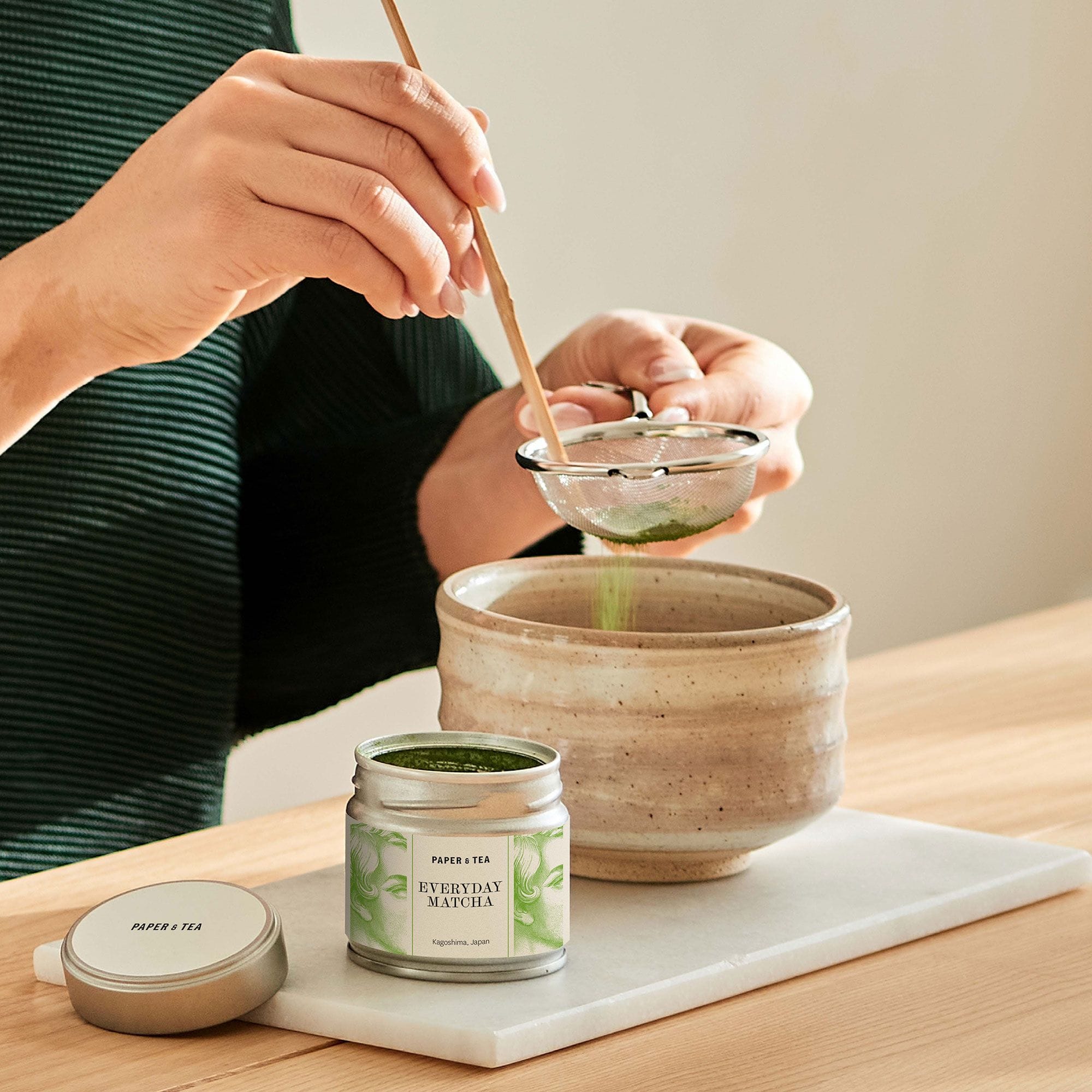 Hände sieben grünes Matcha-Pulver mit Bambusbesen in beige Keramikschale, neben Paper & Tea Matcha-Dose.