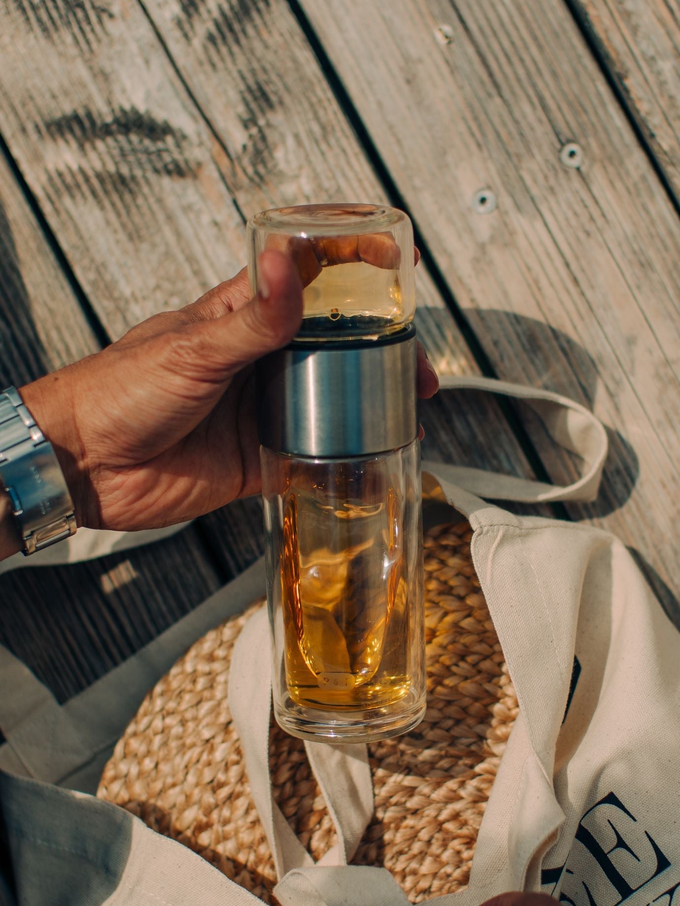 Hand hält Glasflasche mit bernsteinfarbener Flüssigkeit über Segeltuchtasche auf Untersetzer und Holzplanken.