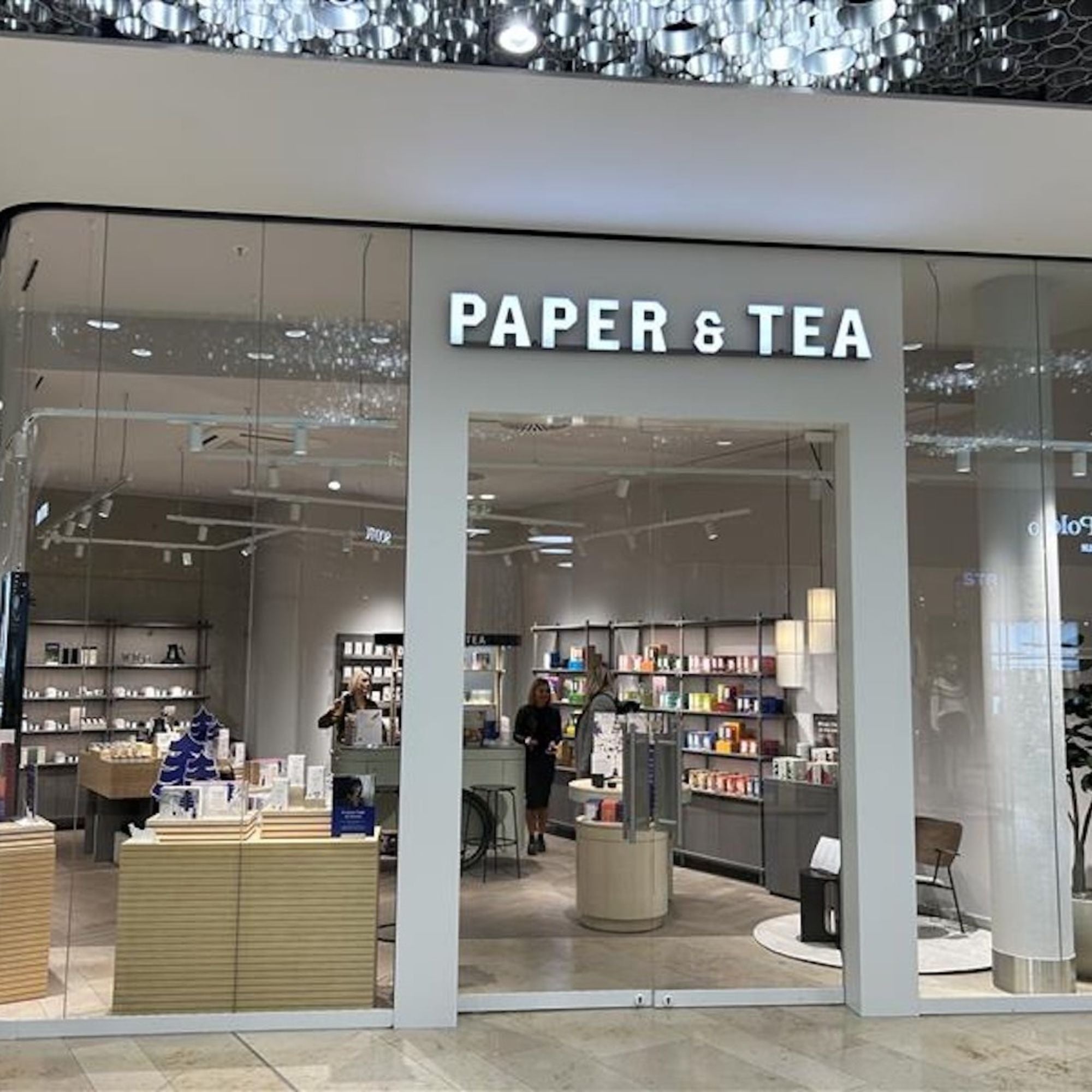Modernes PAPER & TEA Geschäft mit beleuchtetem Eingang und stilvoller Einrichtung.