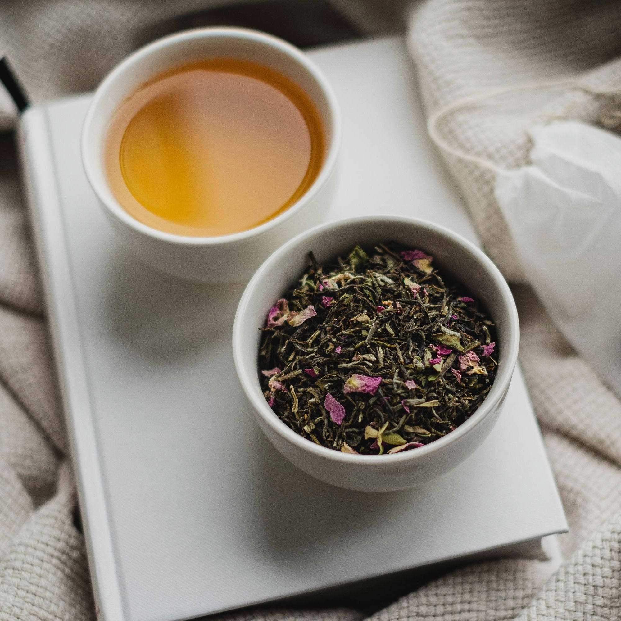 Weißer Keramikbecher mit bernsteinfarbenem Tee und Schale mit losen Teeblättern auf gestricktem Stoff.