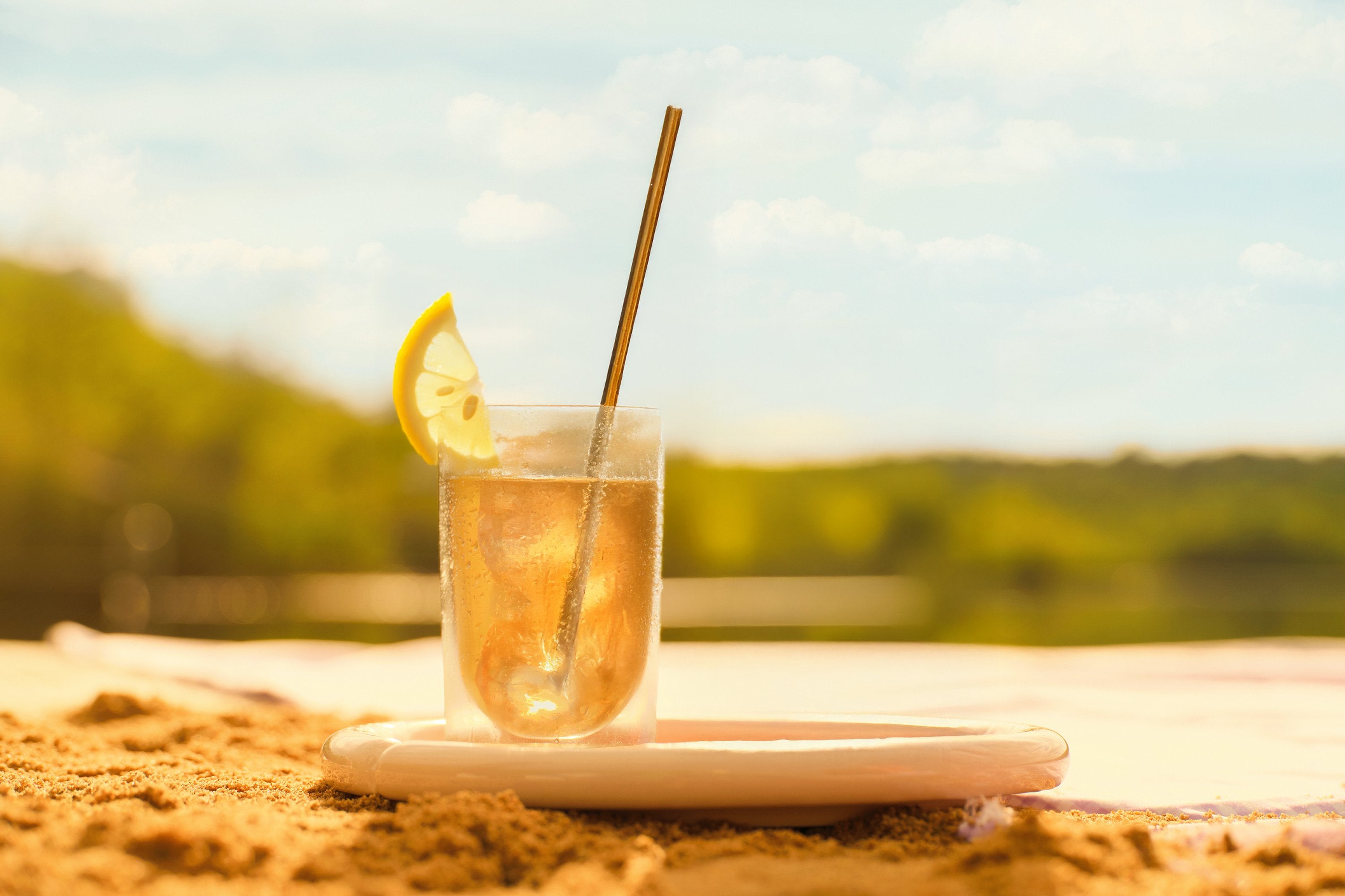 Glas Eistee mit Zitronenscheibe und Metallstrohhalm auf Keramikteller im Sand am See mit grünem Hintergrund