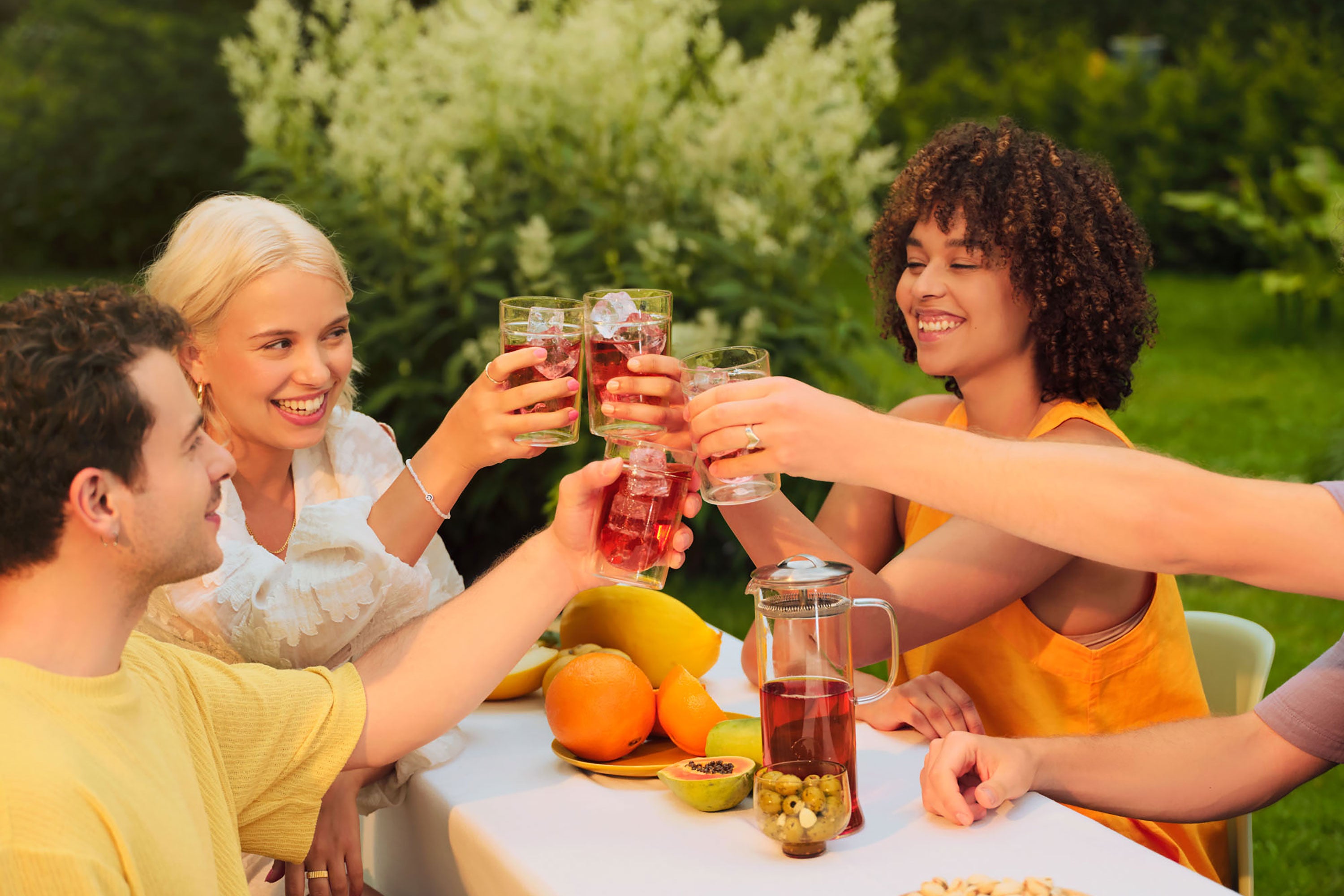 Vier junge Erwachsene stoßen mit roten Eistee-Gläsern an, umgeben von frischen Früchten in einem grünen Garten.