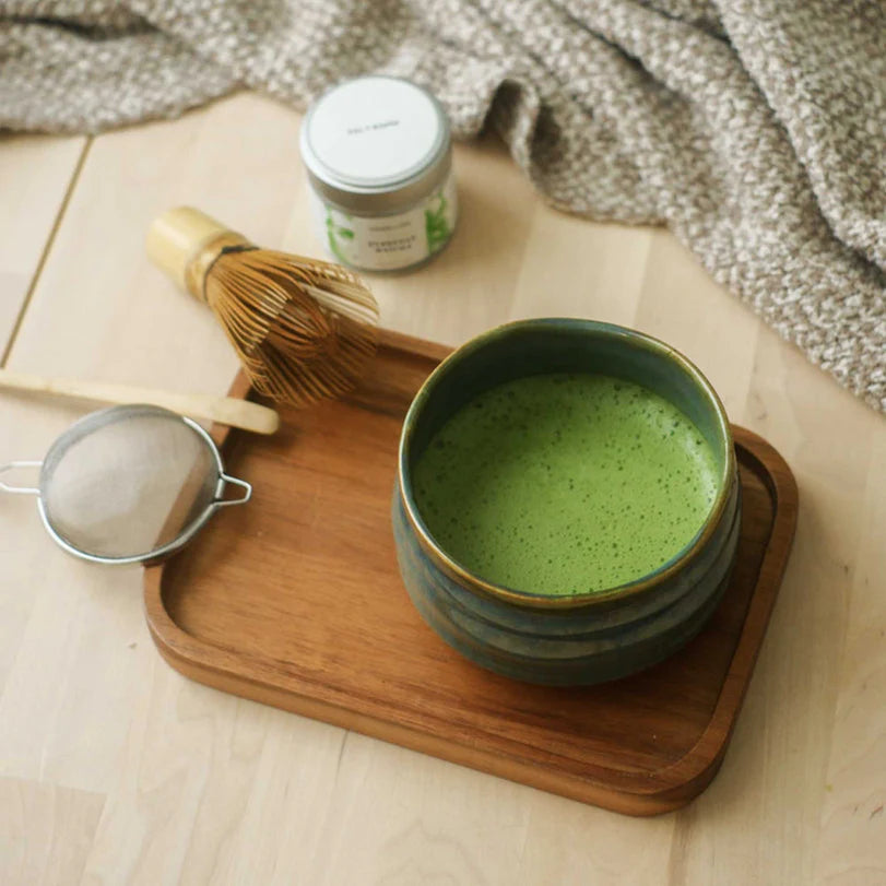 Matcha-Schale mit schaumigem Tee auf Holztablett, umgeben von Bambusbesen, Sieb und Matcha-Behälter.
