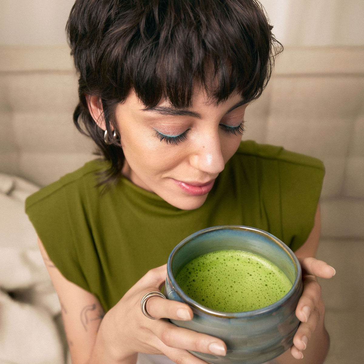 Junge Frau in olivgrünem Top hält Keramiktasse mit Matcha-Tee und lächelt sanft.