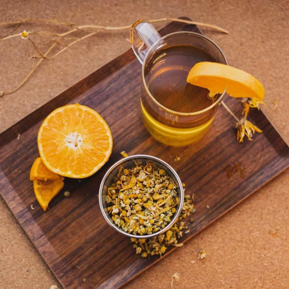 Transparente Glastasse mit hellem Tee und Orangenscheibe auf Holzbrett, umgeben von halber Orange und Kamillenblüten.