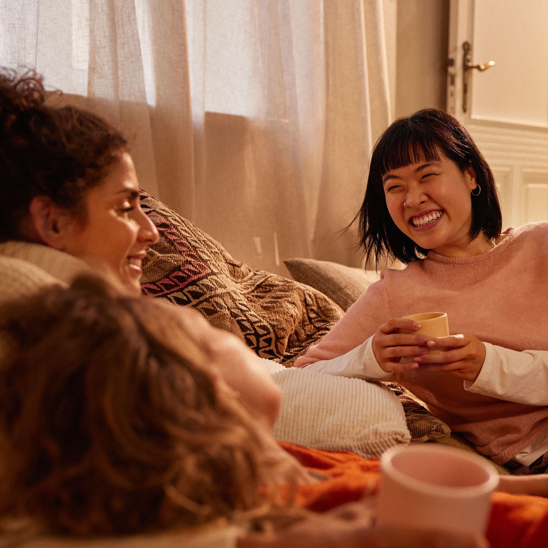 Drei Frauen auf einer Couch, eine lächelt mit gelber Teetasse, beige Vorhänge, gemusterte Kissen.
