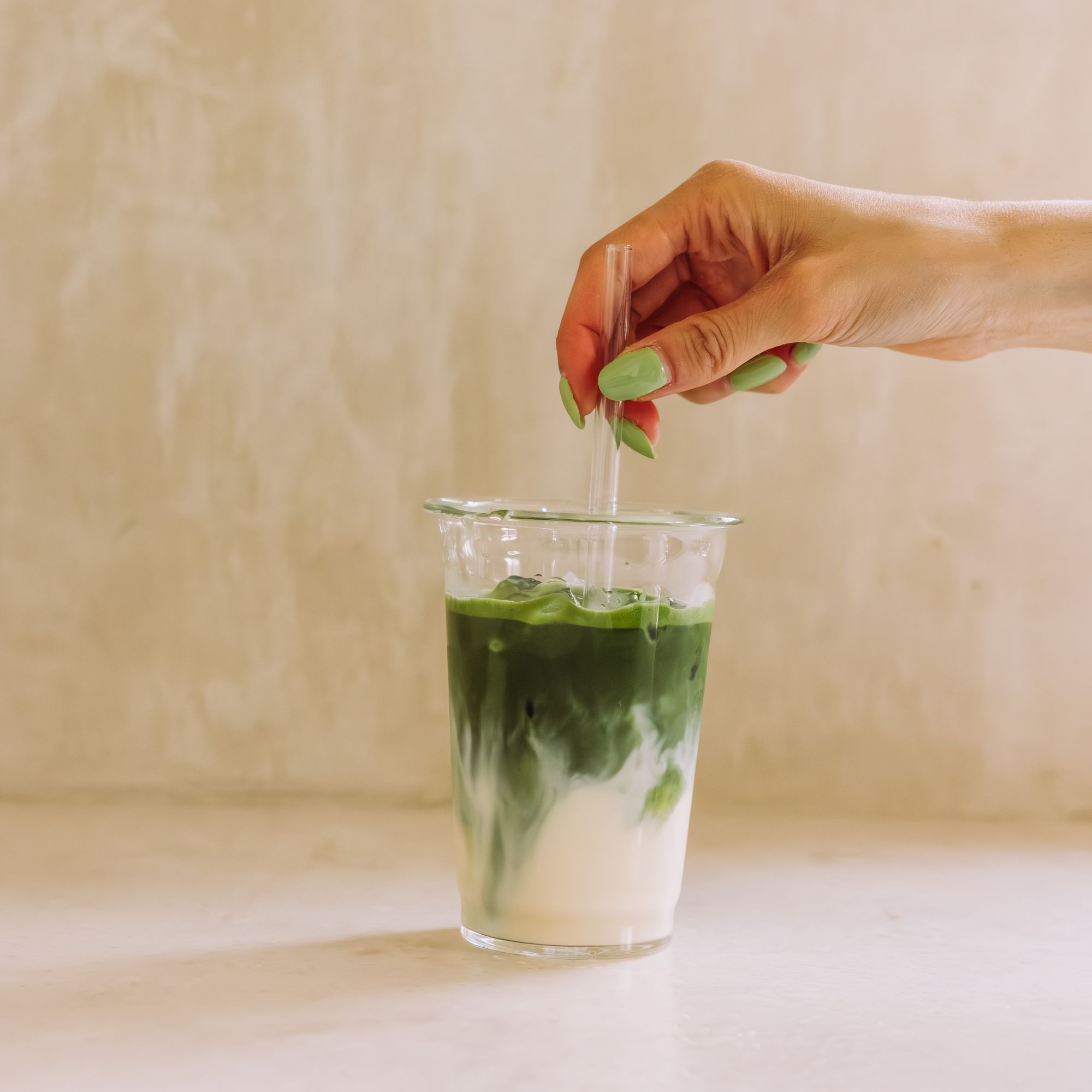 Klare Tasse mit geschichtetem Eistee Latte aus Matcha und Milch, Hand mit grün lackierten Nägeln hält Strohhalm.