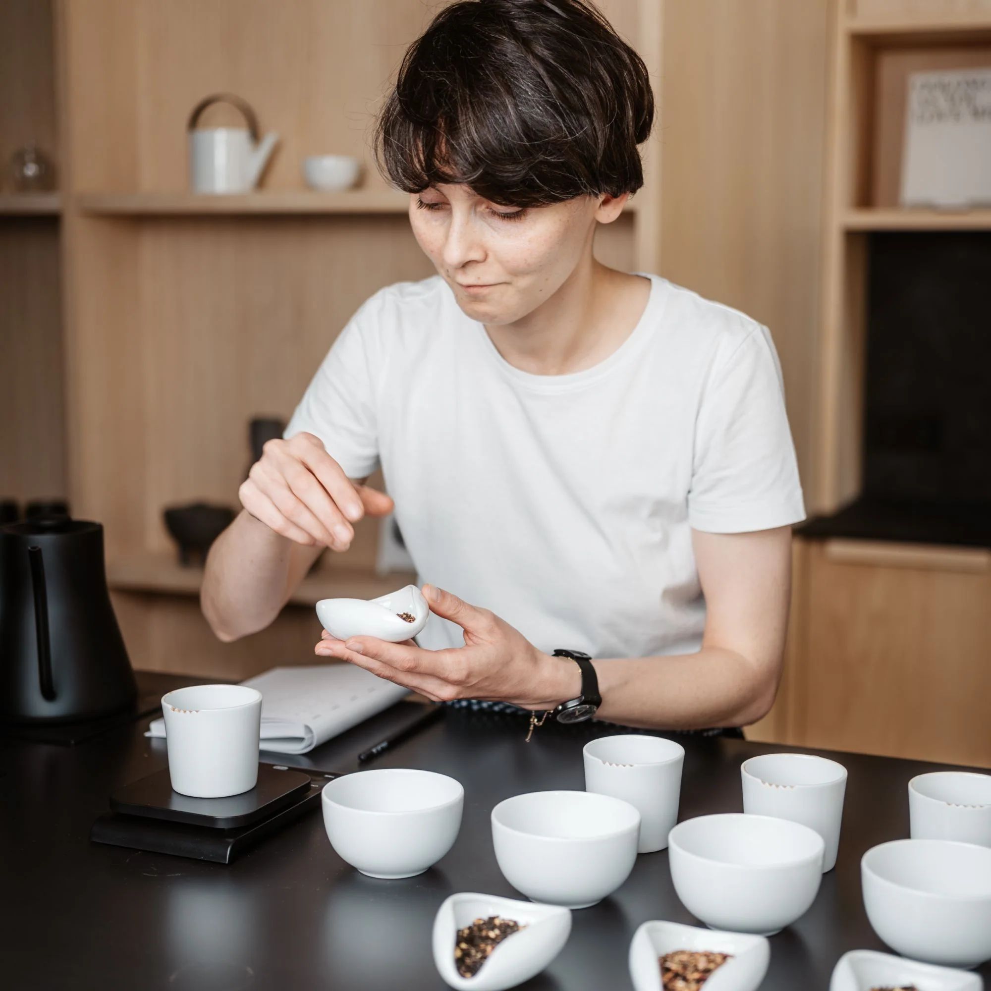Person mit kurzem Haar und weißem T-Shirt untersucht Teeproben in kleinen Schalen auf schwarzem Tisch.