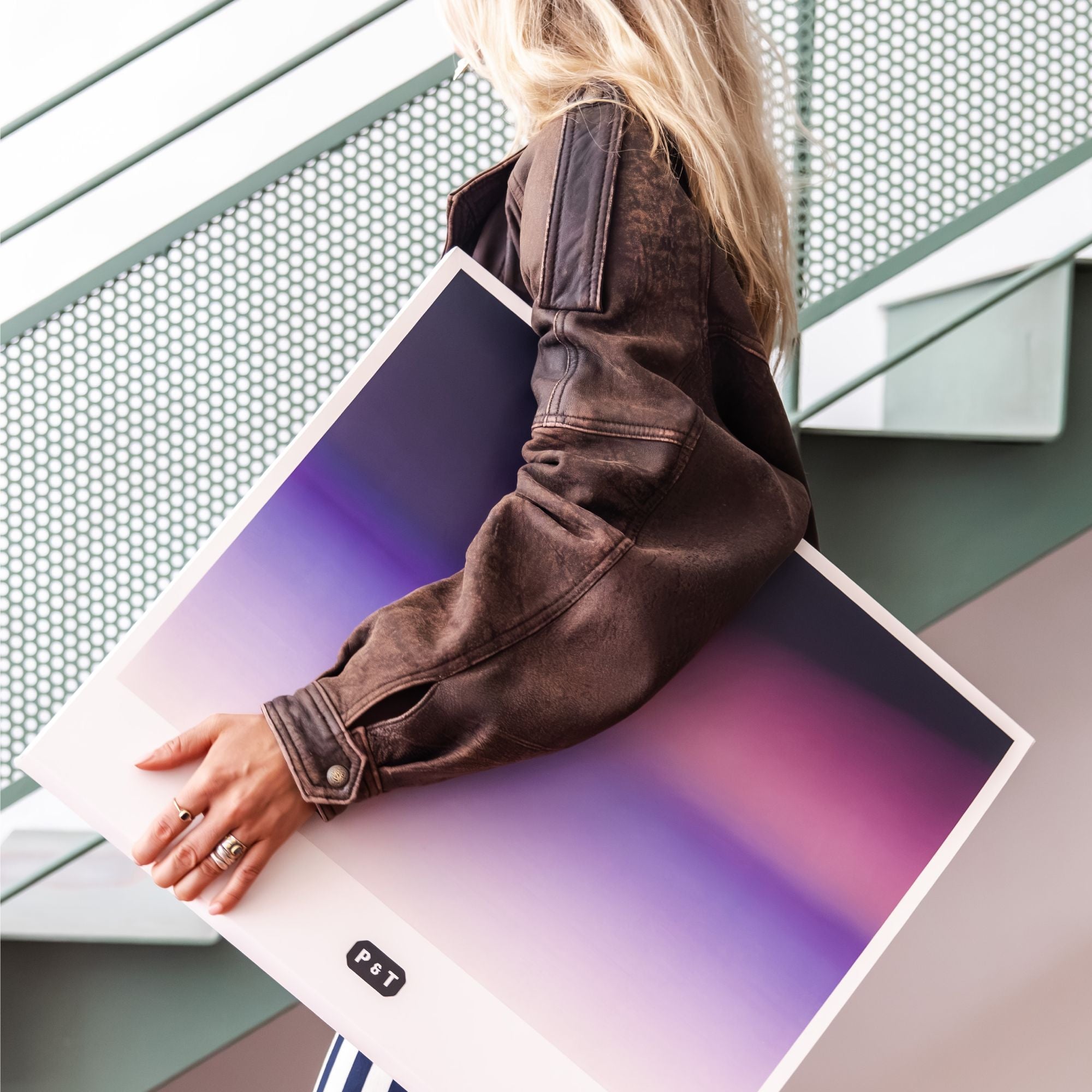 Person in brauner Lederjacke hält Poster mit lila-pinkem Farbverlauf und P&T-Schriftzug vor Metallwand.