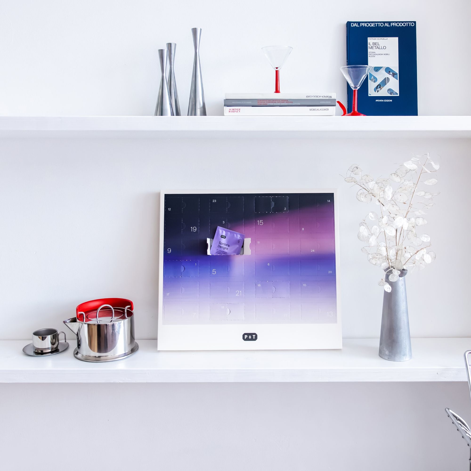 Moderner weißer Wandregal mit blau-lila P&T Adventskalender, metallischem Teekessel mit rotem Griff, Tasse und silberner Vase mit getrockneten weißen Blumen