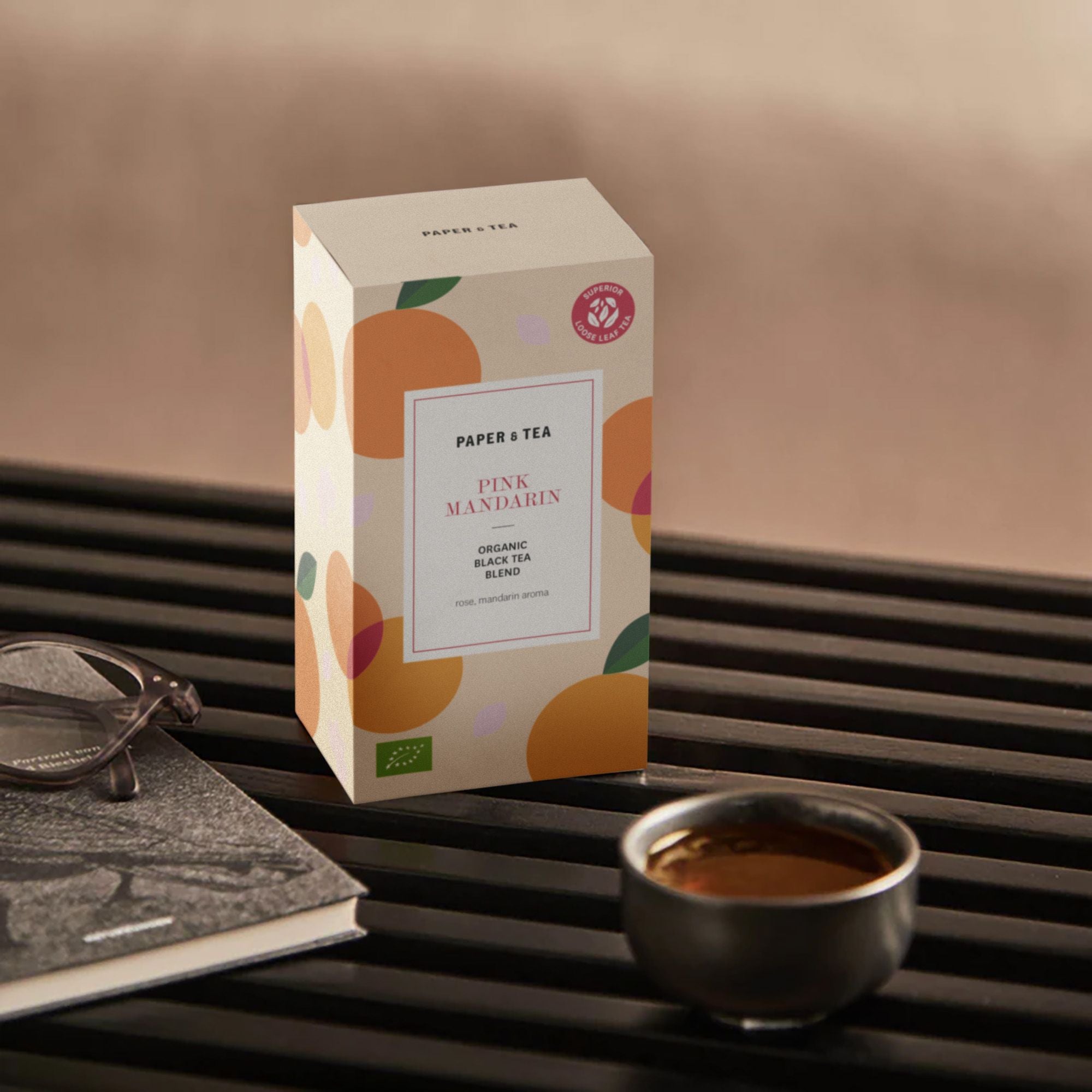 Beige Teebox mit Fruchtmotiven, schwarze Keramiktasse mit Tee, Buch mit Brille auf schwarzer Oberfläche