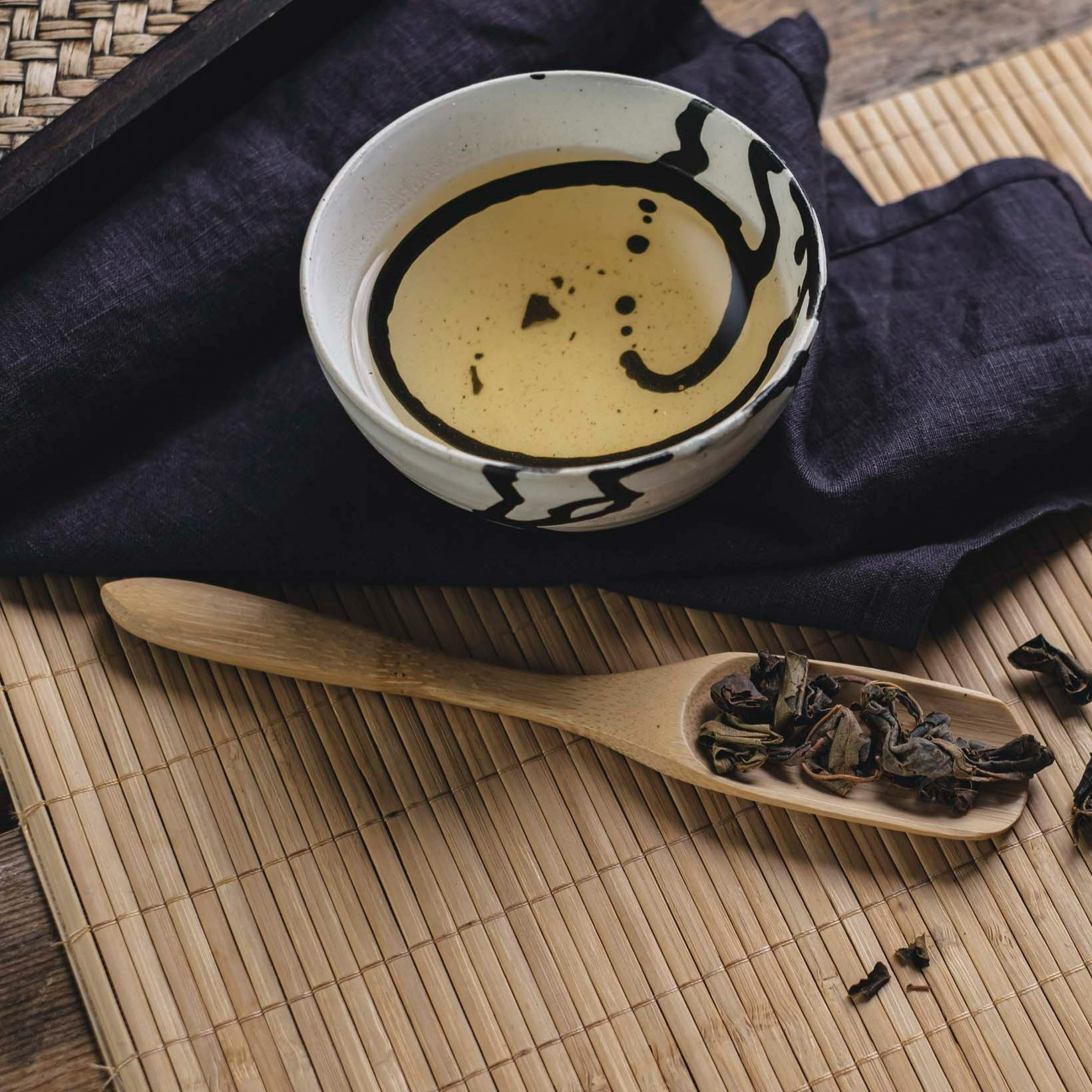 Keramikschale mit schwarzem Muster, gefüllt mit hellem gelben Tee, auf blauem Tuch, neben Holzlöffel mit Teeblättern.