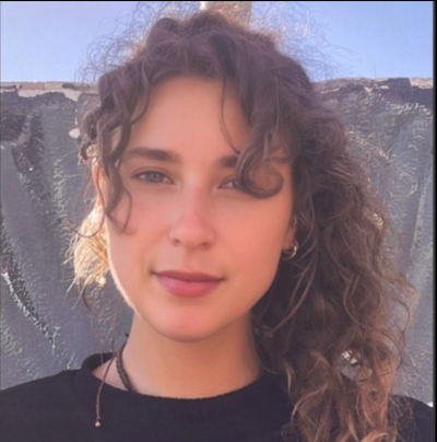 Junge Frau mit lockigem braunem Haar und schwarzem Oberteil vor grauer Putz- und Backsteinwand.