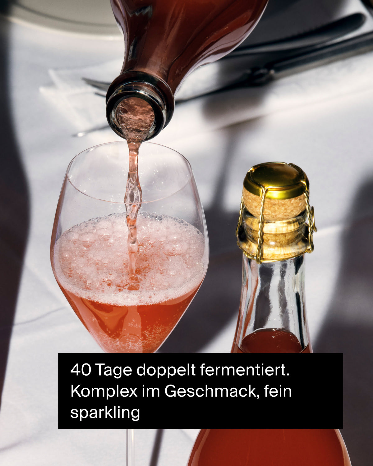 Roséfarbenes, prickelndes Getränk aus brauner Flasche im Weinglas auf weißer Tischdecke, Flasche mit Korken daneben