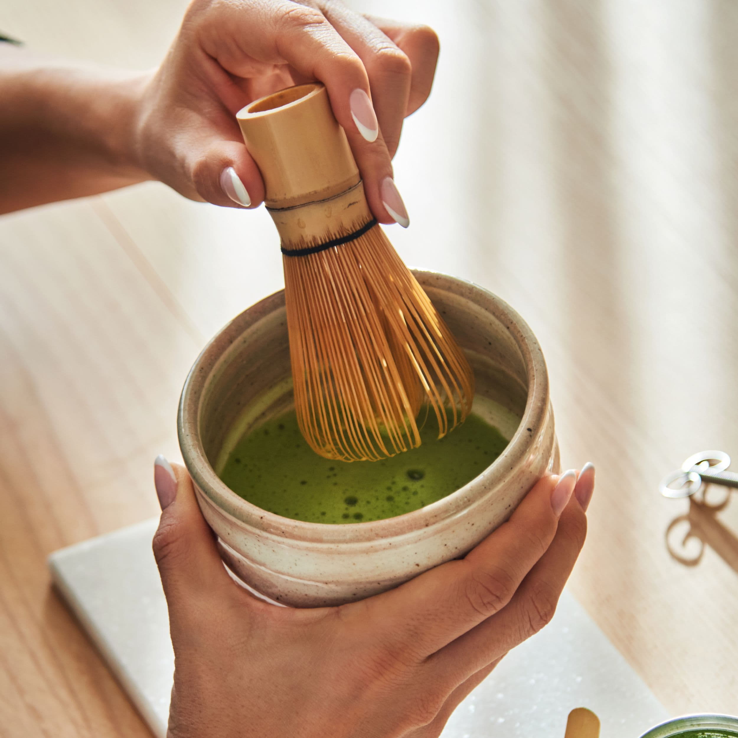 Zwei Hände halten einen Bambusbesen über einer Keramikschale mit schaumigem, grünem Matcha-Tee auf einem Holztisch.