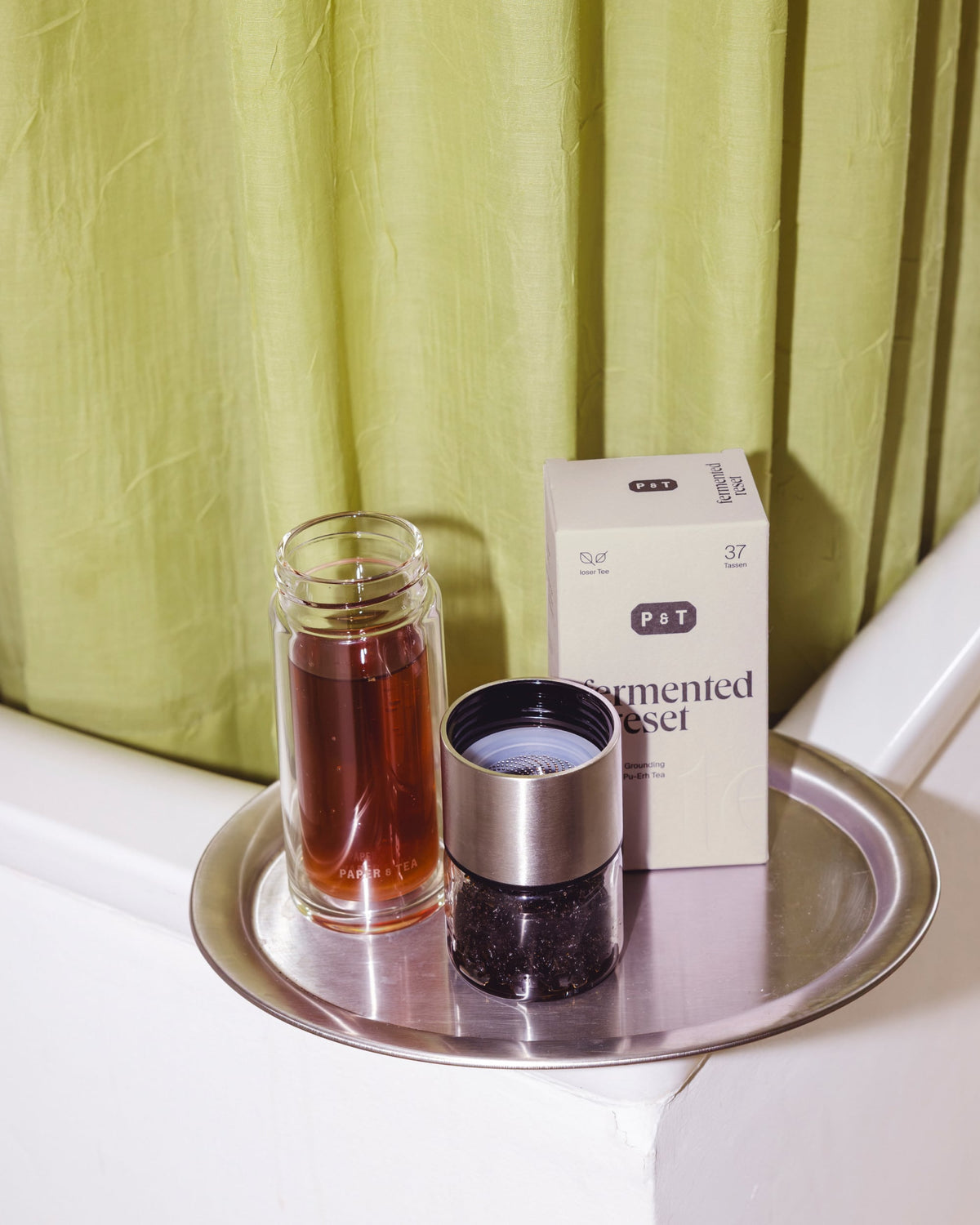 Silbertablett mit Glasgefäß rotem Tee, P&T Teeschachtel 'Grounding Pu-Erh', Metall-Teefilter am Badewannenrand vor grünem Vorhang.