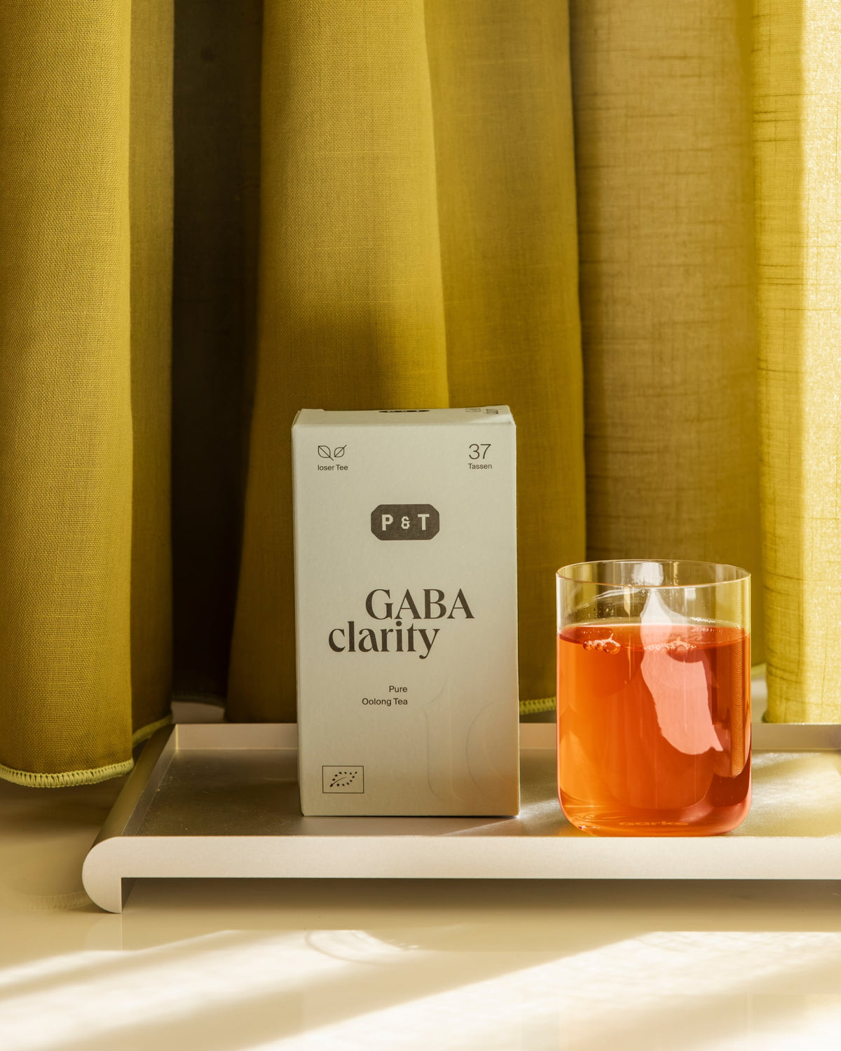 Weiße Verpackung von P&T GABA Clarity Pure Oolong Tee neben klarem Glas mit orangefarbenem Aufguss auf Tablett.