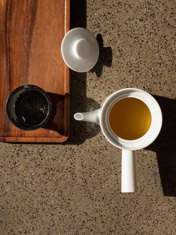 Weißer Keramik-Teekessel und heller grüner Tee in schwarzem Gefäß auf Holztablett vor Steinoberfläche