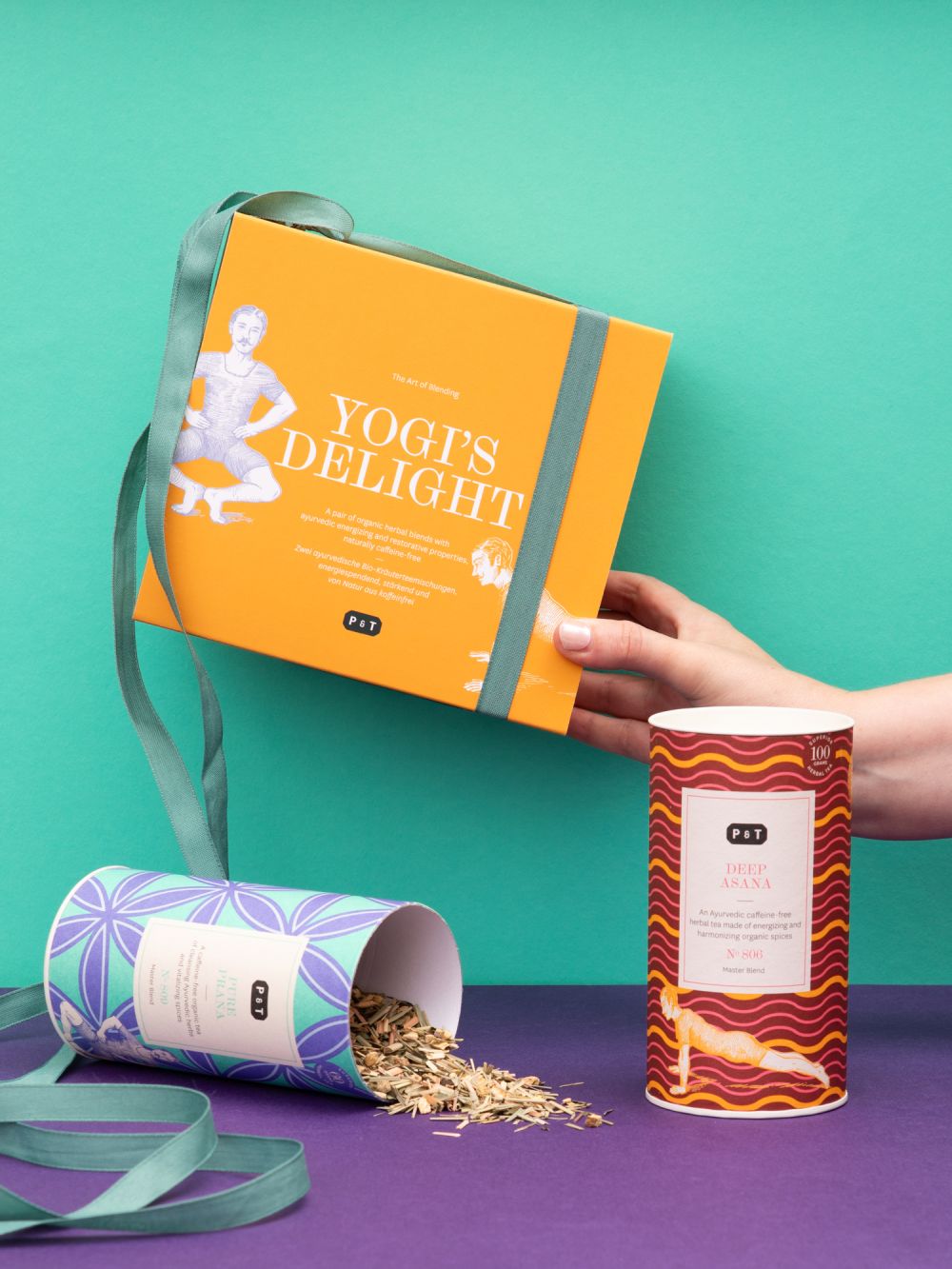 Leuchtend orange Yogi's Delight Teebox und rote Deep Asana Teedose vor türkis-lila Hintergrund.