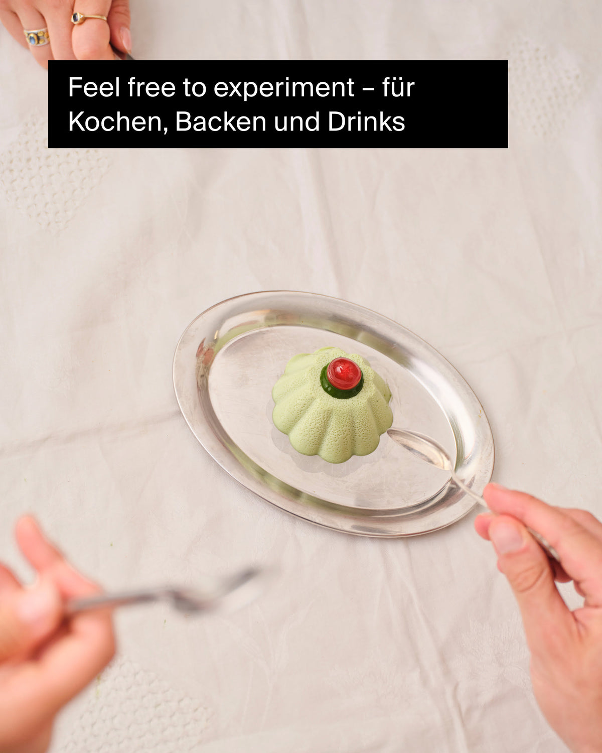 Grünes Gelee mit roter Garnitur auf silbernem Tablett, zwei Hände mit Löffeln bereit zum Genießen.