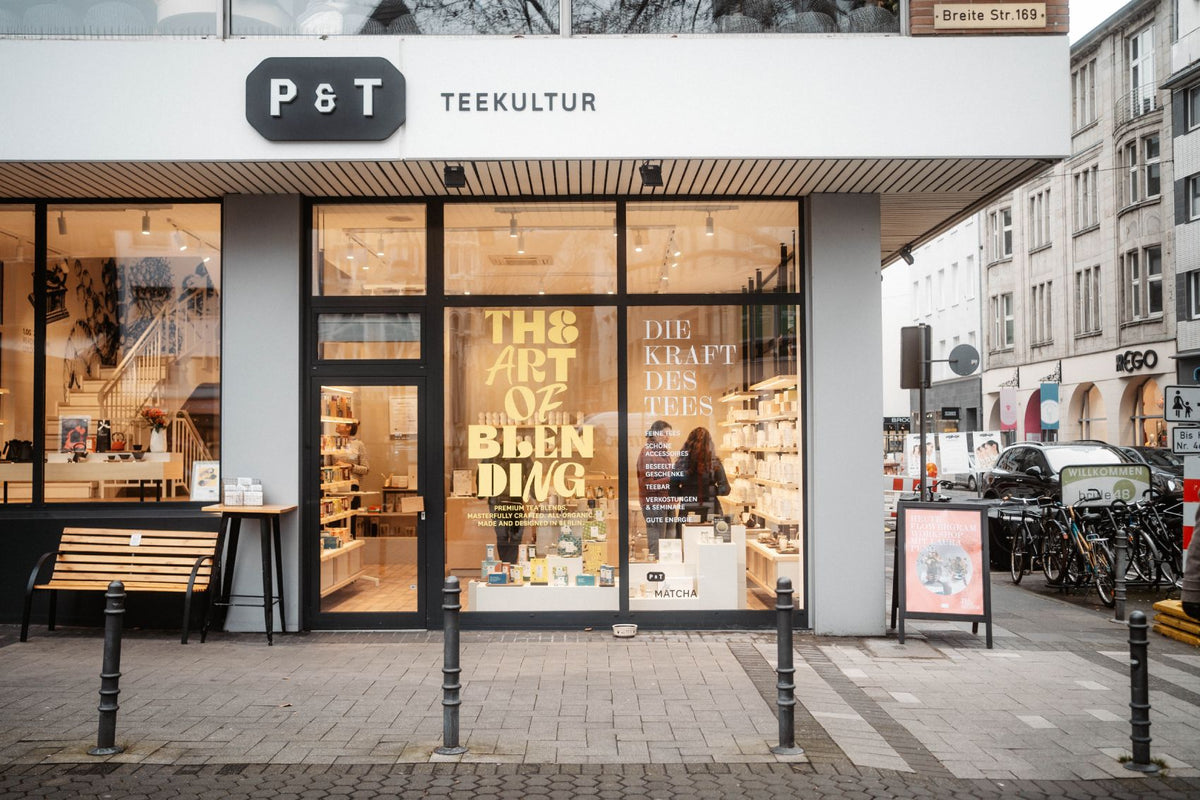 P&T Teekultur-Ladenfront mit weißem Schild, goldener Schrift, Holzbank und Workshop-Banner