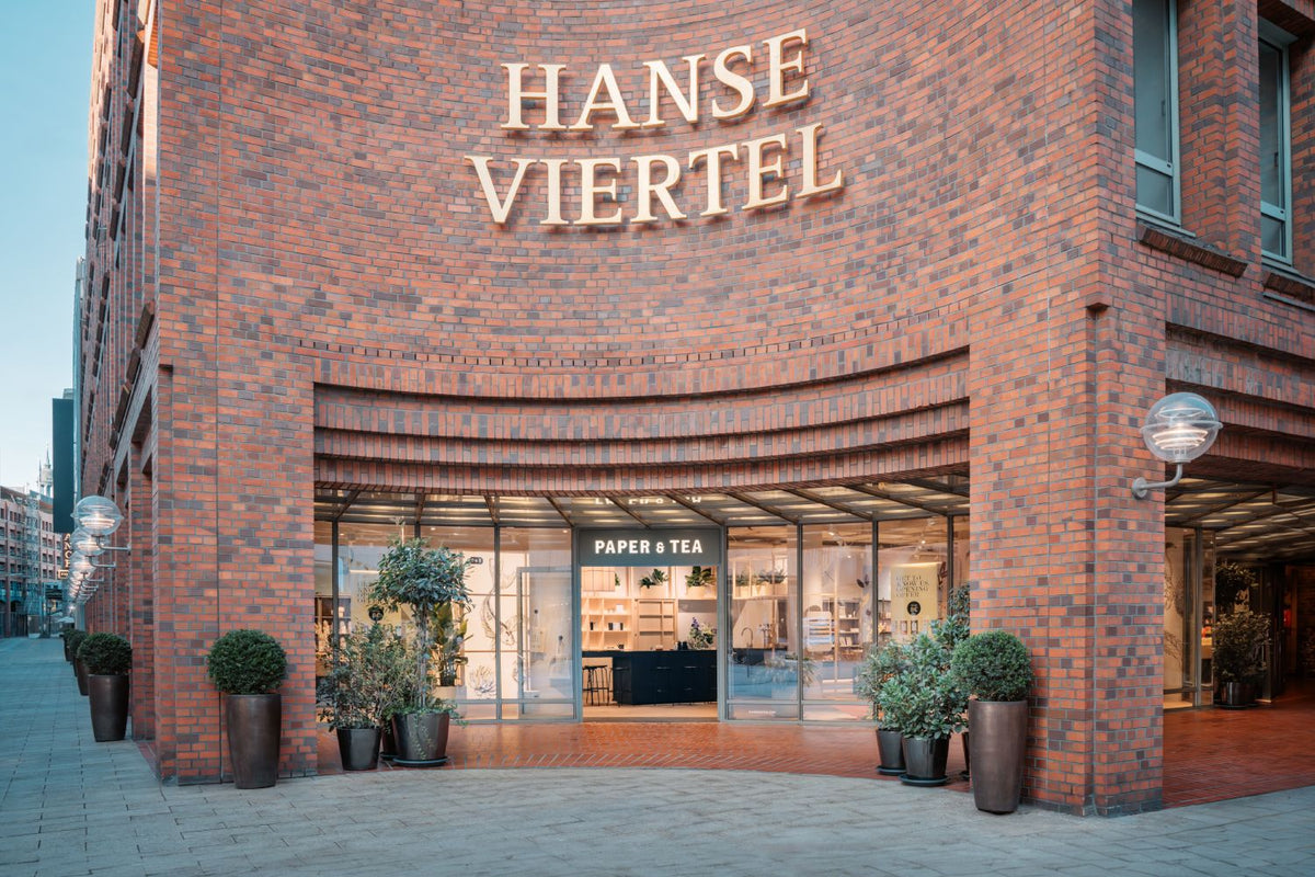 Geschwungene Backsteinfassade mit HANSE VIERTEL Schriftzug und P&T Ladeneingang mit Glasfront und Pflanzen.