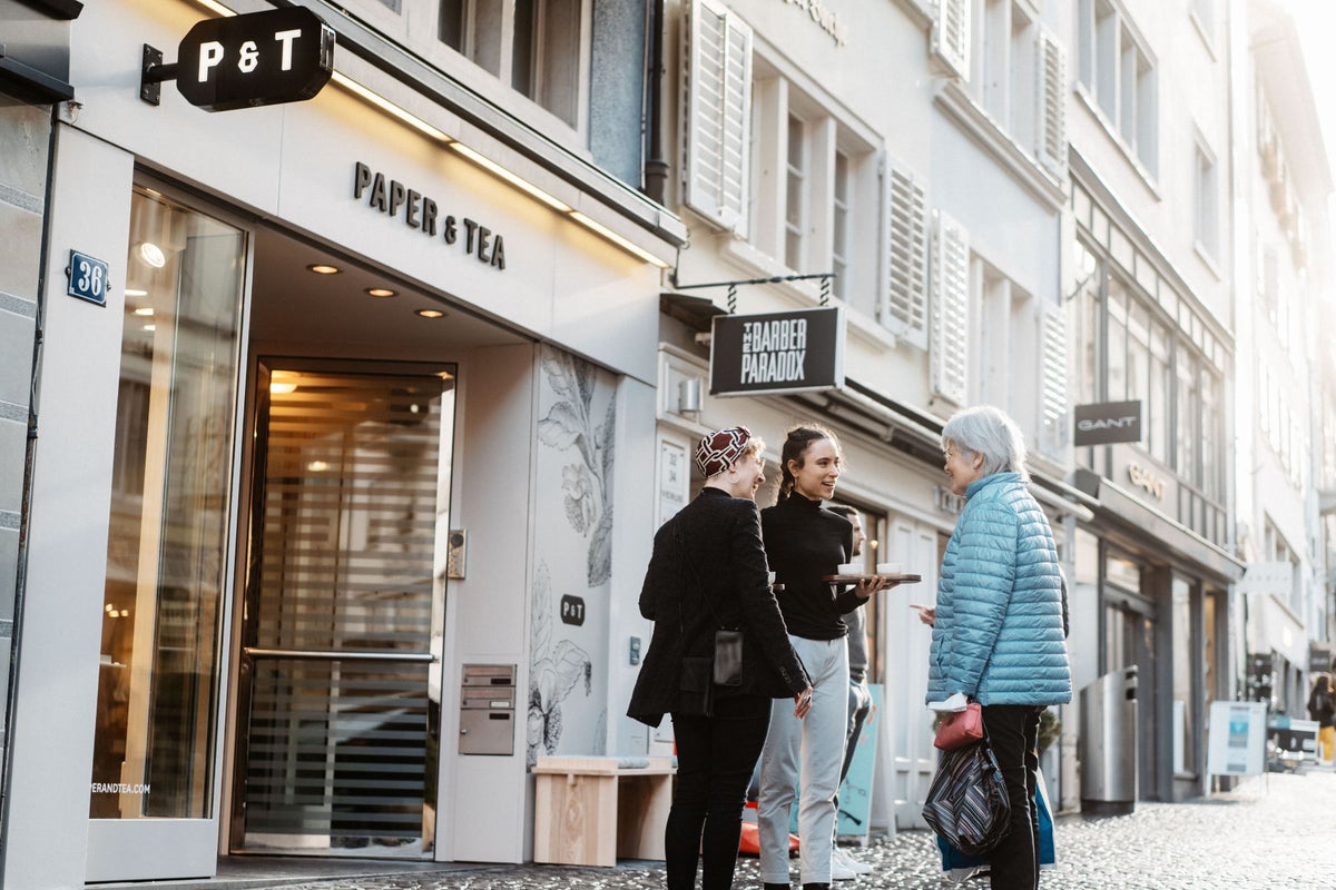 Helle Straßenszene mit P&T Shop-Eingang Nr. 36, drei Frauen im Gespräch mit Teetassen auf einem Tablett