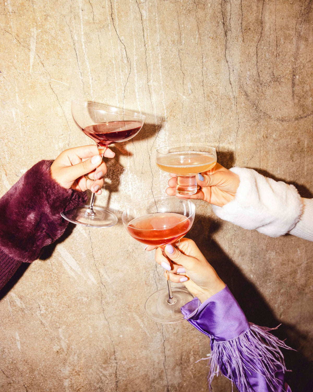 Drei Hände mit farbenfrohen Cocktailgläsern in eleganten, texturierten Ärmeln vor beiger Wand.