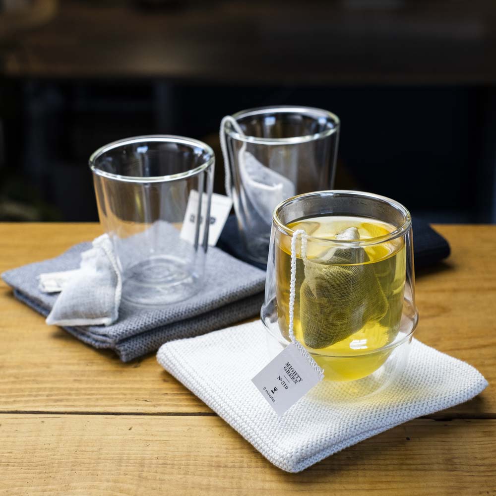 Drei doppelwandige Gläser auf Holztisch mit Mighty Green N-319 Teebeuteln und ziehendem Grüntee auf weißem Tuch.
