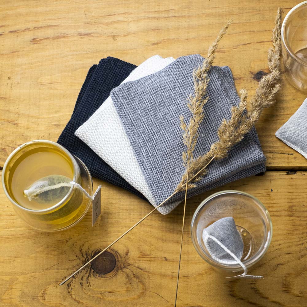 Drei gefaltete gestrickte Tücher in Dunkelblau, Weiß und Hellgrau auf Holztisch mit Tee und Weizenhalmen