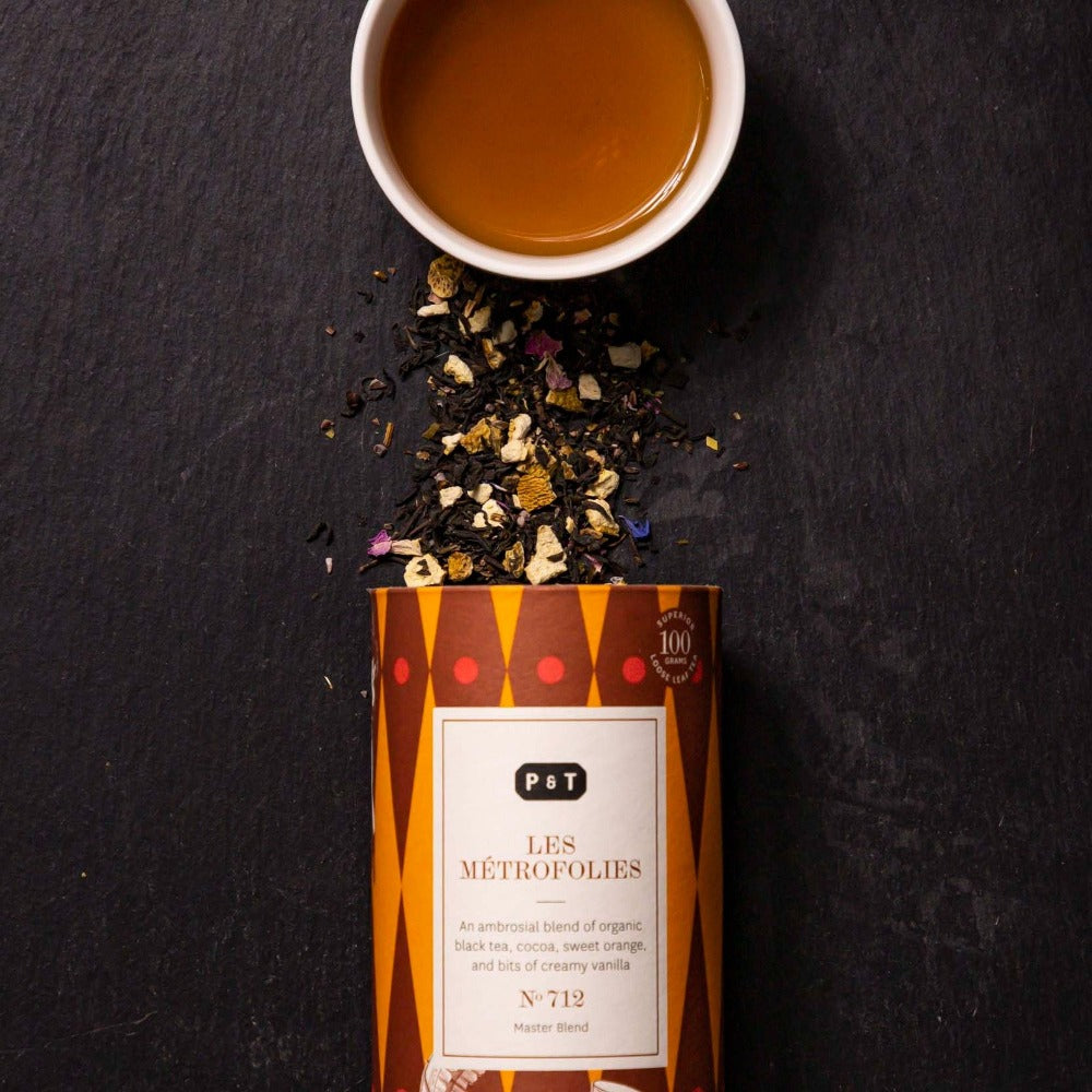 Dose mit LES MÉTROFOLIES Bio-Schwarztee, Tasse Bernstein-Tee und lose Teeblätter auf dunklem Hintergrund.