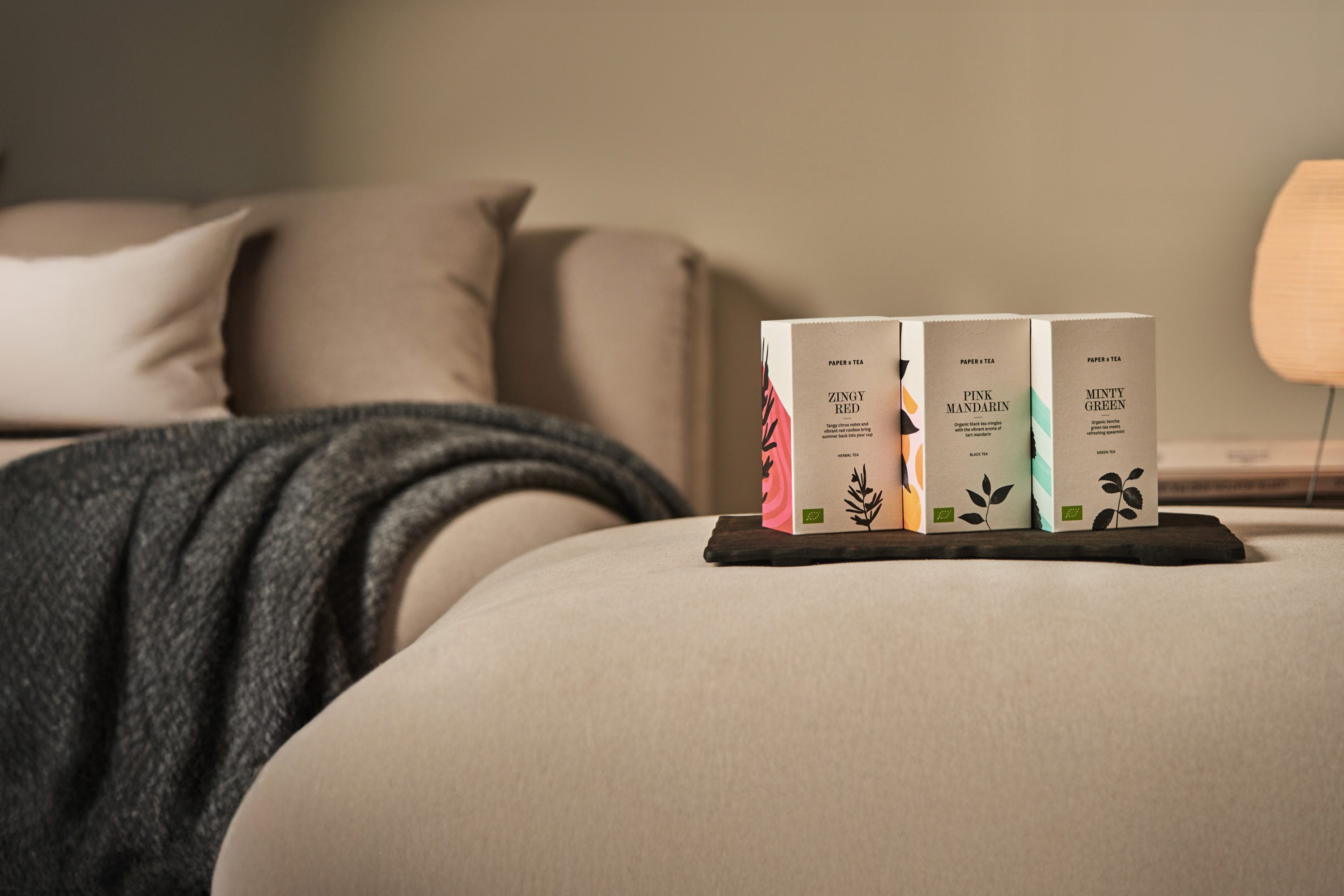 Drei PAPER & TEA Teeboxen – Zingy Red, Pink Mandarin und Minty Green – auf schwarzem Tablett vor Sofa.