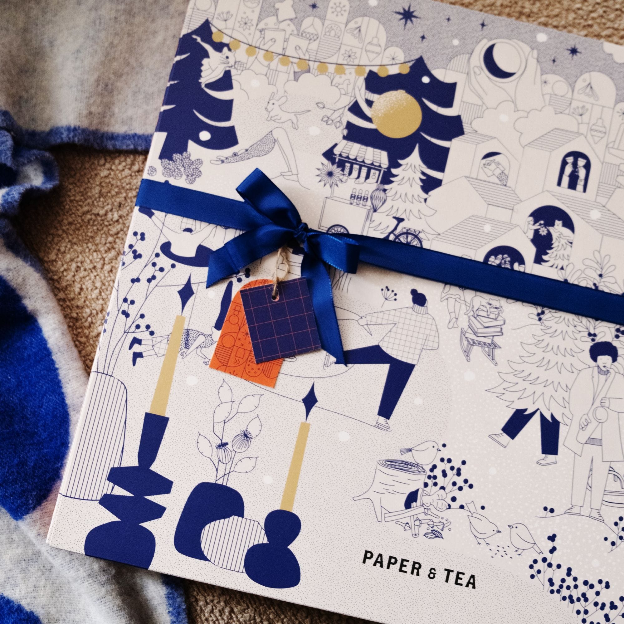 Winterliche PAPER & TEA Geschenkbox mit blau-weißen Illustrationen und blauem Satinband auf Teppich.