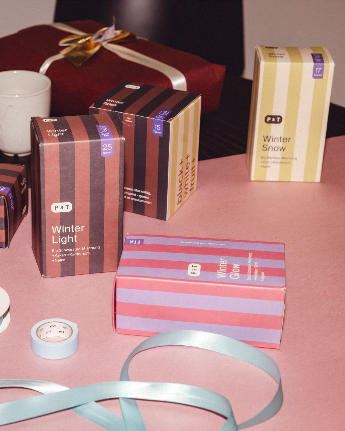 Vier farbige Winter-Teepakete von P&T mit Bio-Mischungen, weiße Tasse und rotes Geschenk mit goldener Schleife.
