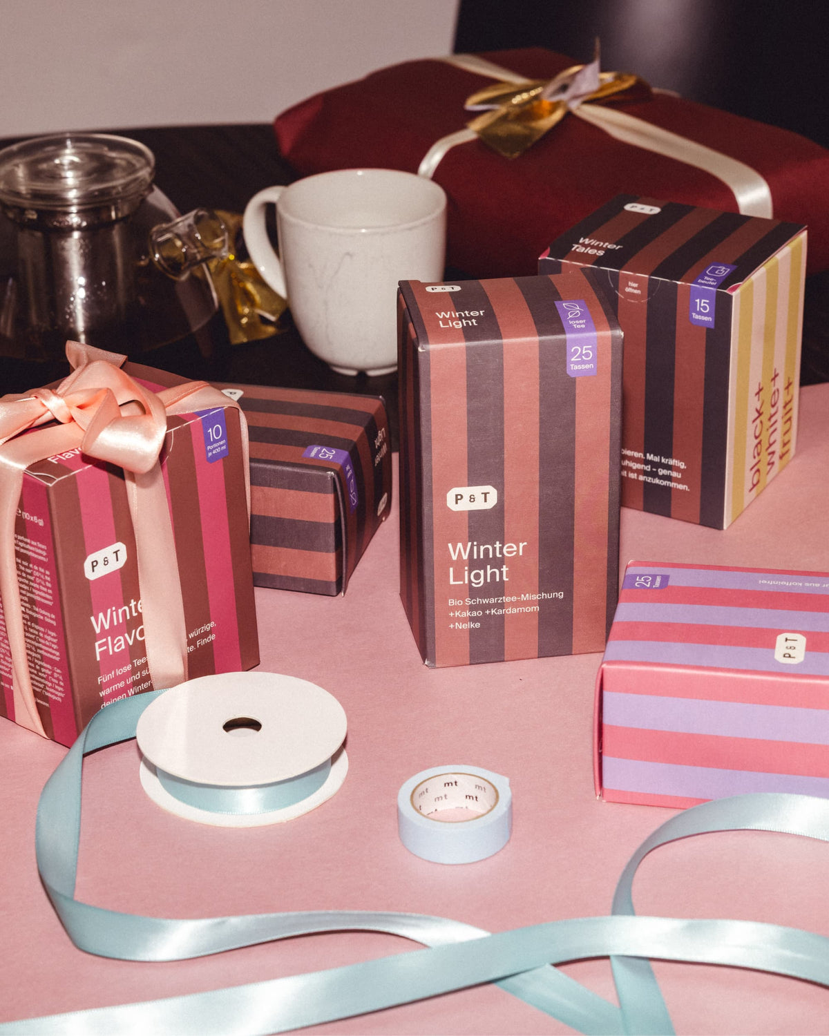 Bunte P&T-Teeboxen Winter Light und Winter Tales, Keramiktasse und Teekanne auf rosa Tisch mit blauer Deko.